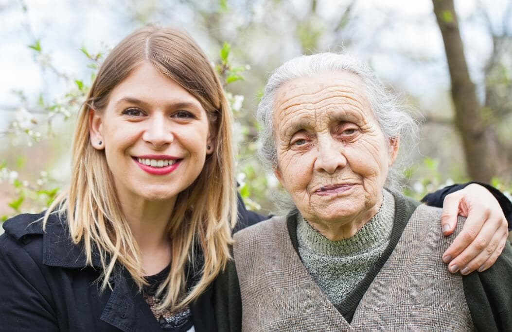 A smiling young woman with her arm around an elderly woman, both standing outdoors with blurred greenery in the background. - Home Instead