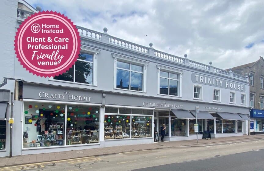 Street view of a white building named "Trinity House" with various shops including "The Crafty Hobbit. - Home Instead