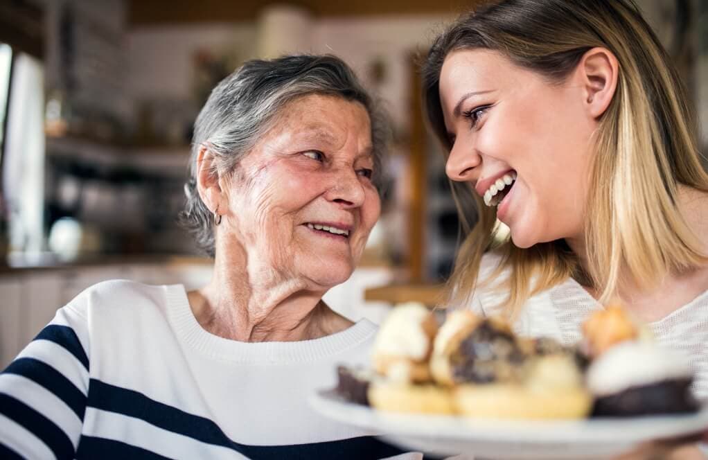 An elderly woman and a young woman smile at each other, holding a plate with assorted pastries. - Home Instead
