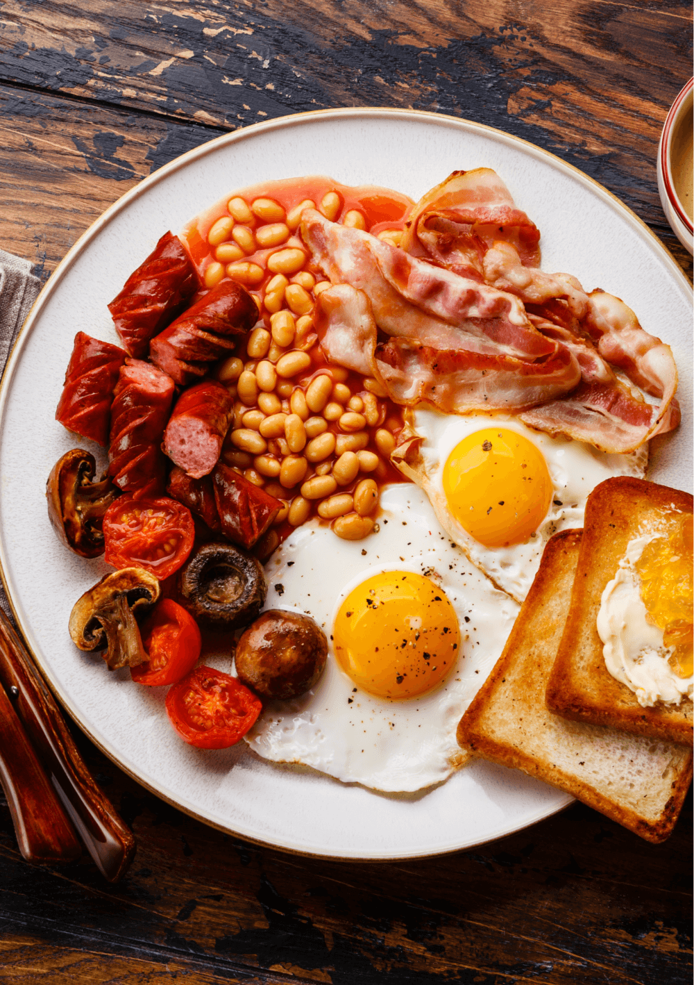 A plate with sunny-side-up eggs, bacon, sausage, baked beans, mushrooms, tomatoes, and toast on a wooden table. - Home Instead