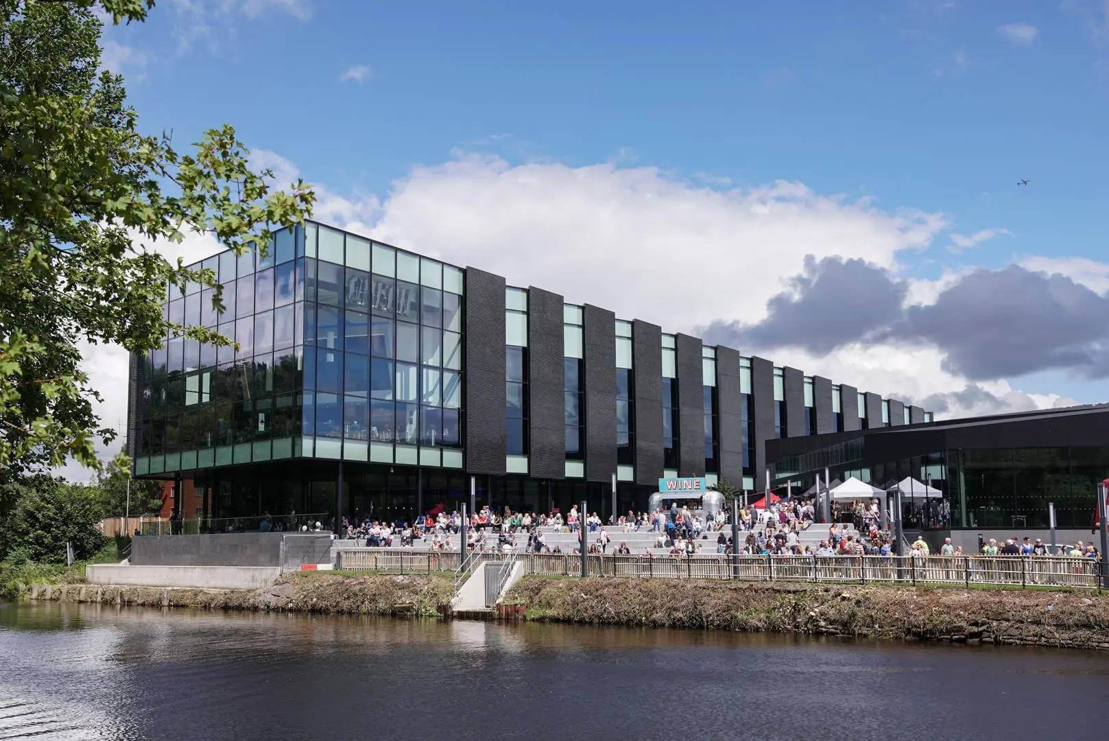 Large modern glass building by a river, with people gathered outside, under a partly cloudy blue sky. - Home Instead