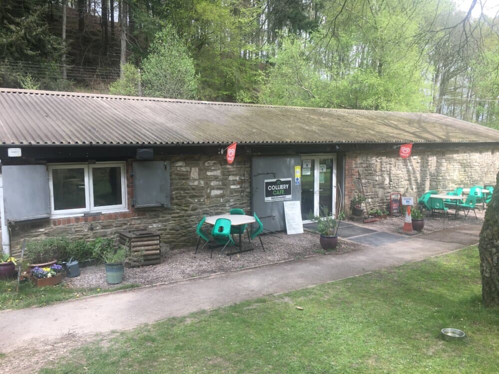 A stone building with green outdoor tables and chairs under trees, featuring a sign that says "Colliery Café. - Home Instead