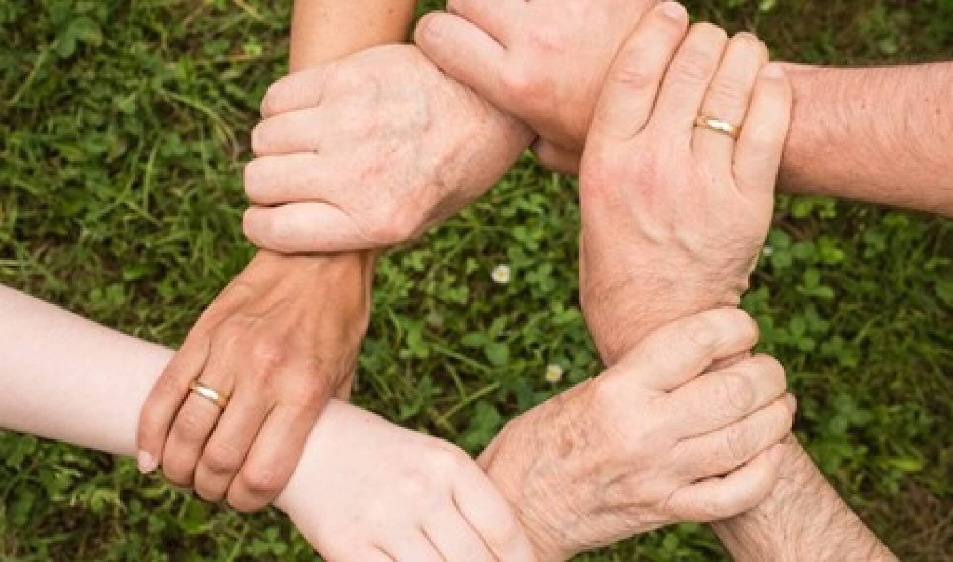 Six hands of different skin tones clasped together in a circle over green grass, symbolizing unity and teamwork. - Home Instead