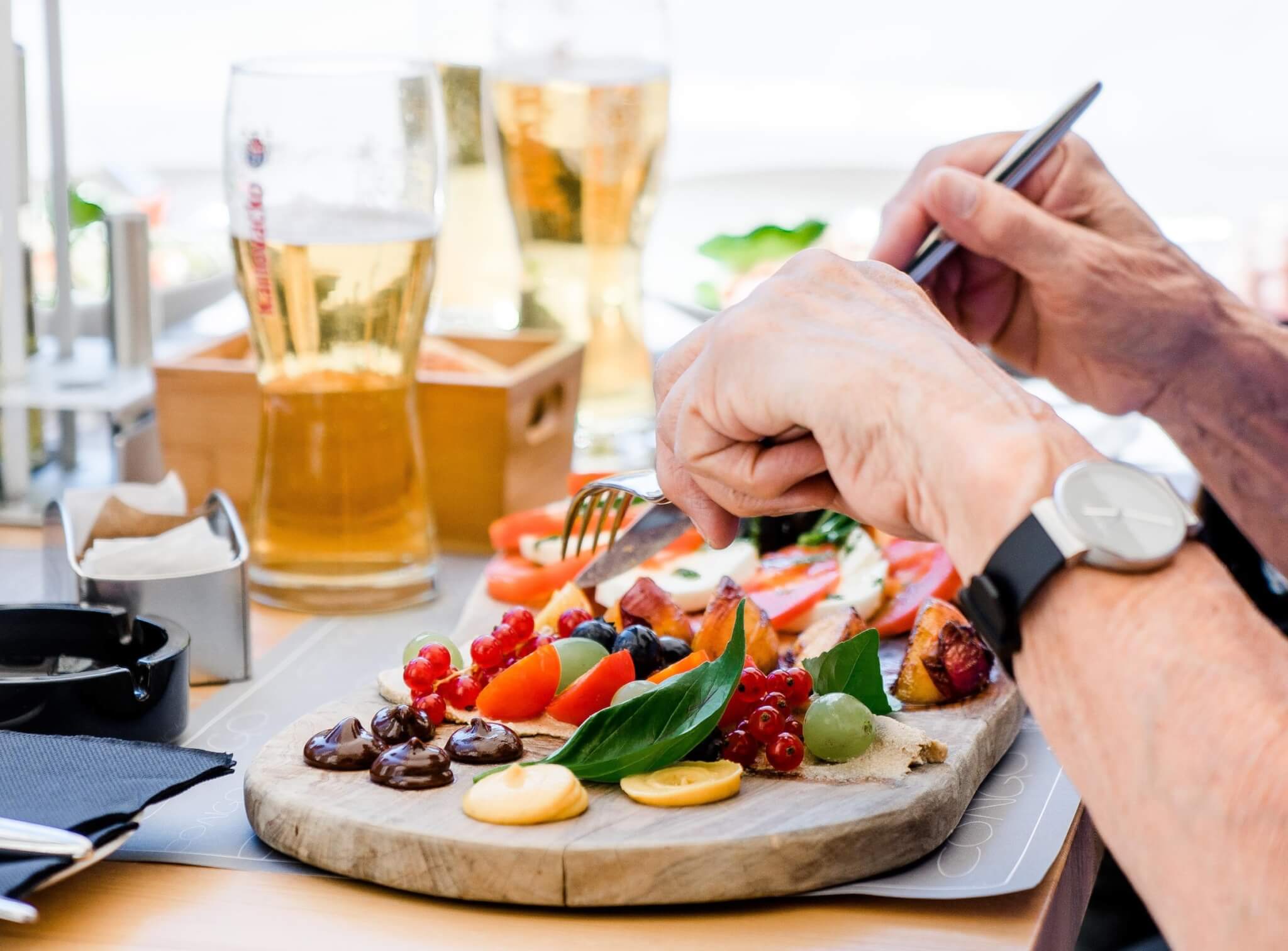Close-up of a person eating a colorful dish on a wooden board with a variety of foods and glasses of beer in the background. - Home Instead