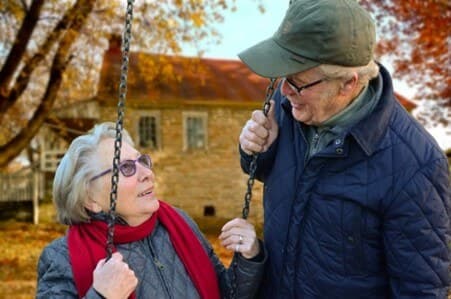 Older couple smiling at each other by a swing in a park with autumn leaves and a brick house in the background. - Home Instead