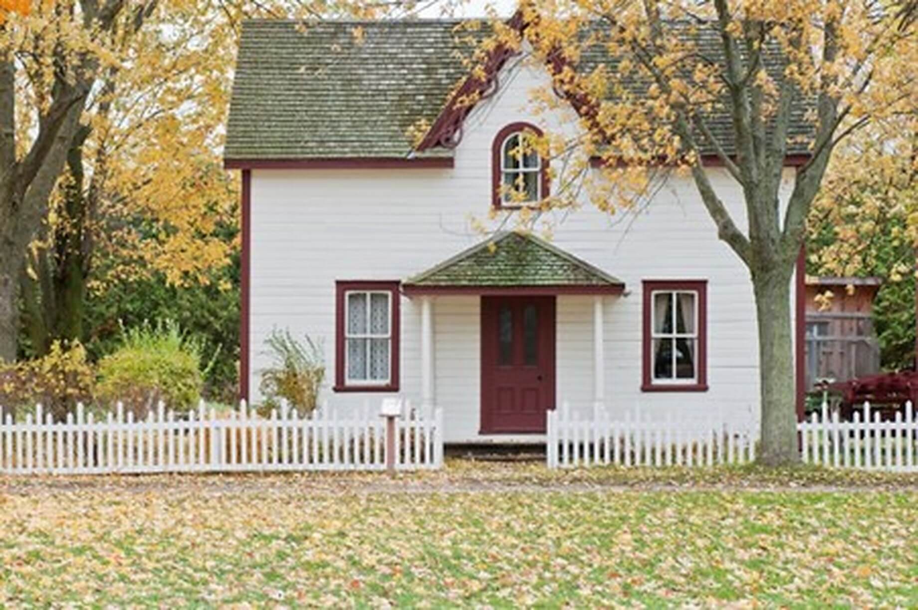 A quaint white house with red trim, surrounded by autumn trees and a white picket fence. Fallen leaves cover the lawn. - Home Instead