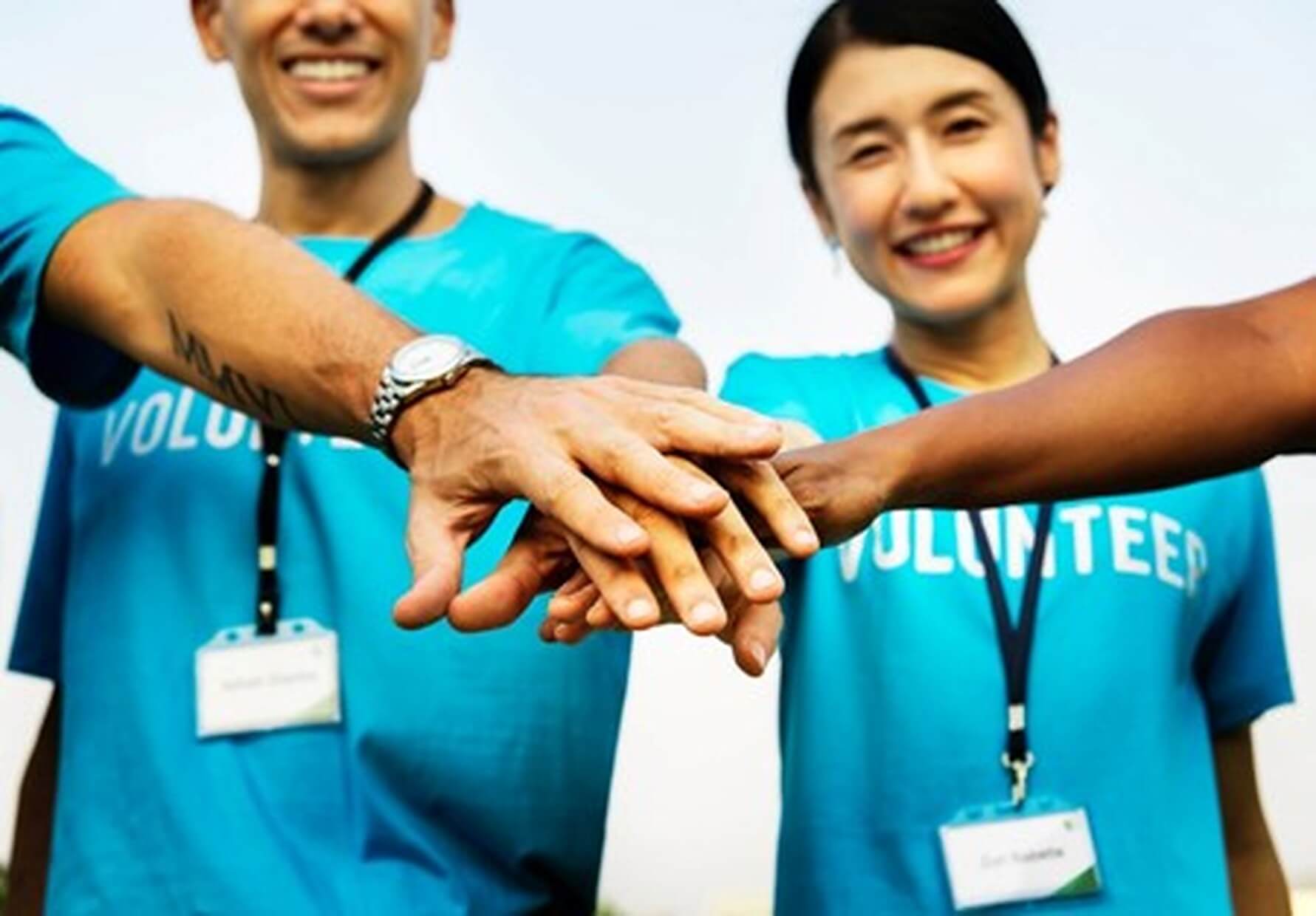 People in blue 'volunteer' t-shirts stacking hands together in unity, smiling and wearing name tags. - Home Instead