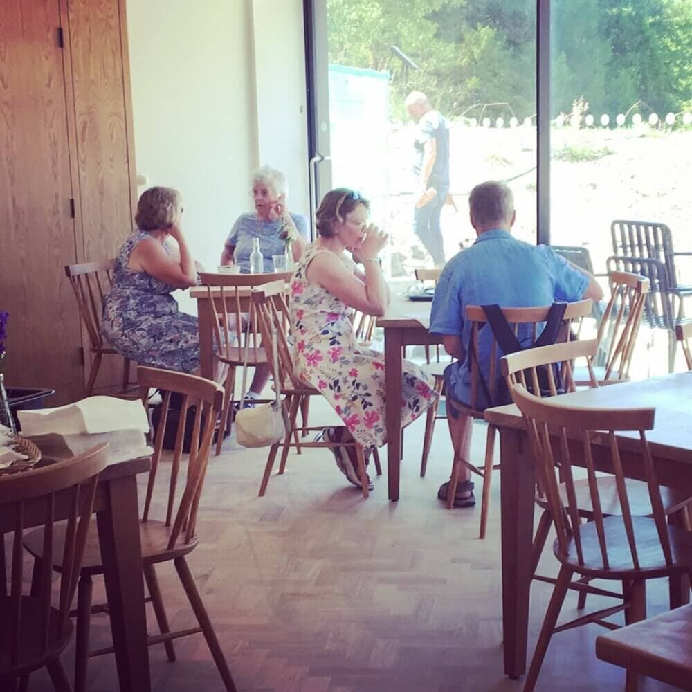 People seated at wooden tables in a sunlit cafe with large windows and wooden chairs. - Home Instead