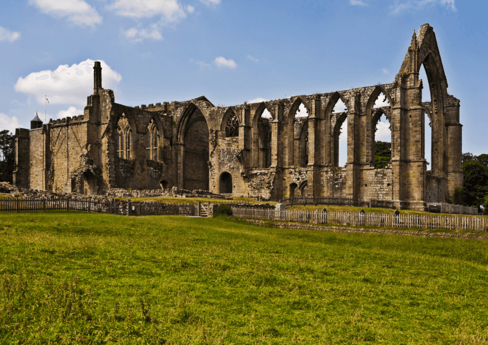 Ancient stone ruins of a partially intact abbey with arches and columns, surrounded by a grassy field under a blue sky. - Home Instead