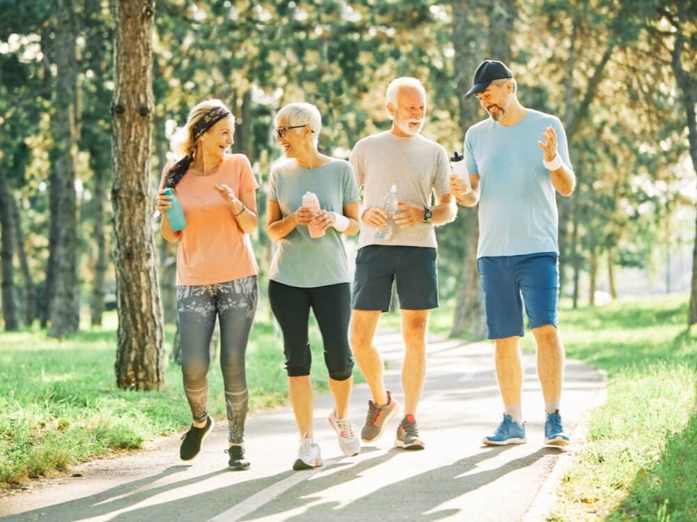 Four adults walking and chatting on a park pathway, holding water bottles and dressed in casual active-wear. - Home Instead