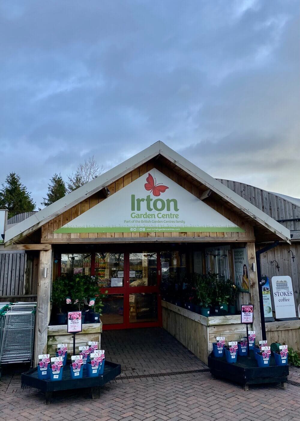 Entrance of Irton Garden Centre with potted plants displayed outside and a sign for Stooks Café. - Home Instead