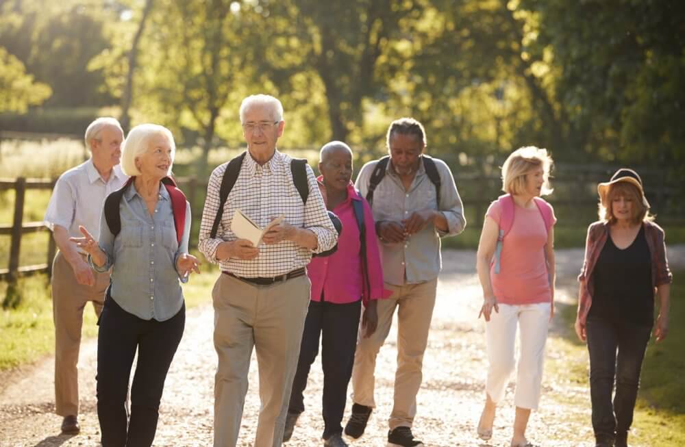 A group of older adults walking outdoors on a sunny day, wearing backpacks and engaging in conversation. - Home Instead