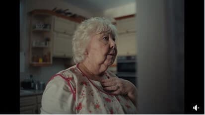 Elderly woman in a floral apron holding her chest, looking contemplative, standing in a dimly lit kitchen. - Home Instead