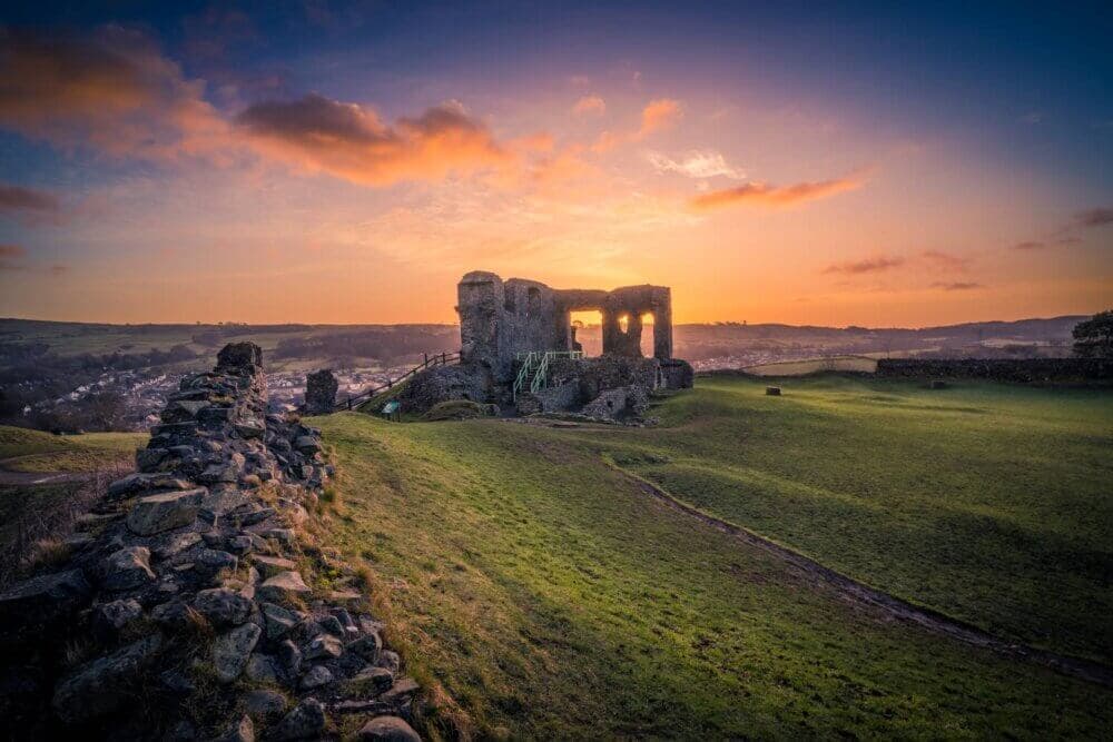 Ancient ruins at sunset, with a stone wall leading towards a crumbling structure on a grassy hill. - Home Instead