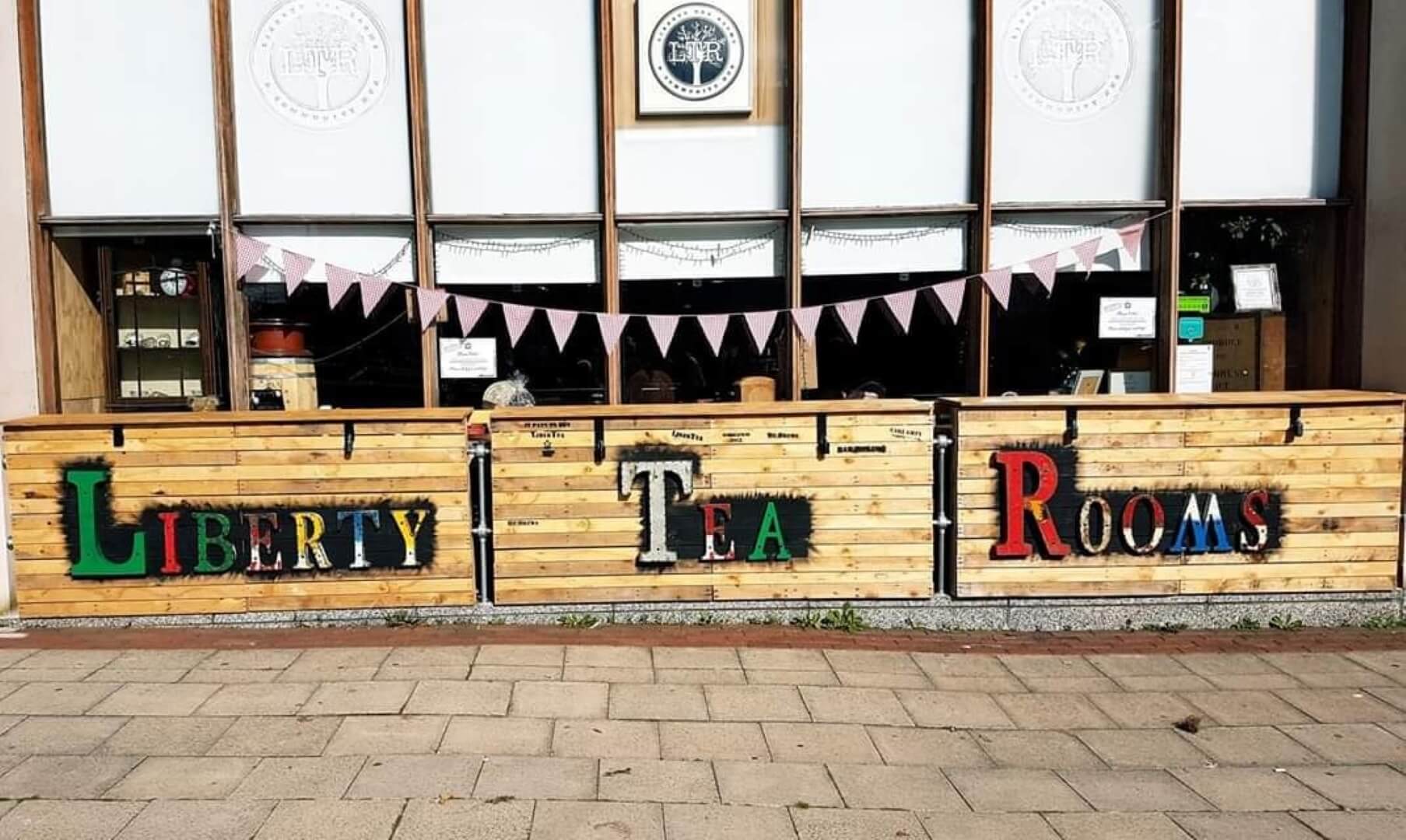 Front of a cafe with "Liberty Tea Rooms" sign and bunting, featuring large windows and a wooden outdoor area. - Home Instead