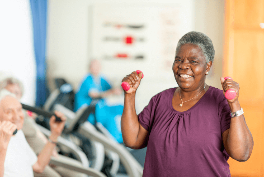 An older woman smiles while lifting small pink weights in an exercise class, with other participants in the background. - Home Instead
