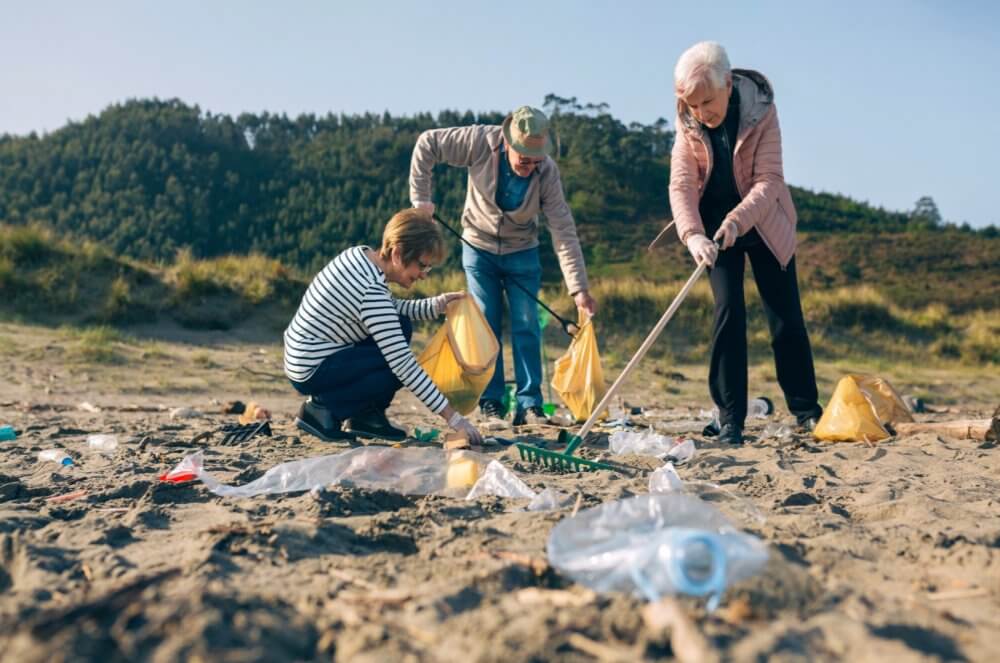 Three people collect trash on a beach, using garbage bags and pickers, with grassy hills in the background. - Home Instead