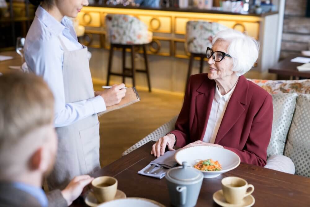 Elderly woman ordering food from a waitress at a restaurant. A man is also seated at the table with tea and pastries. - Home Instead Bournemouth & Christchurch