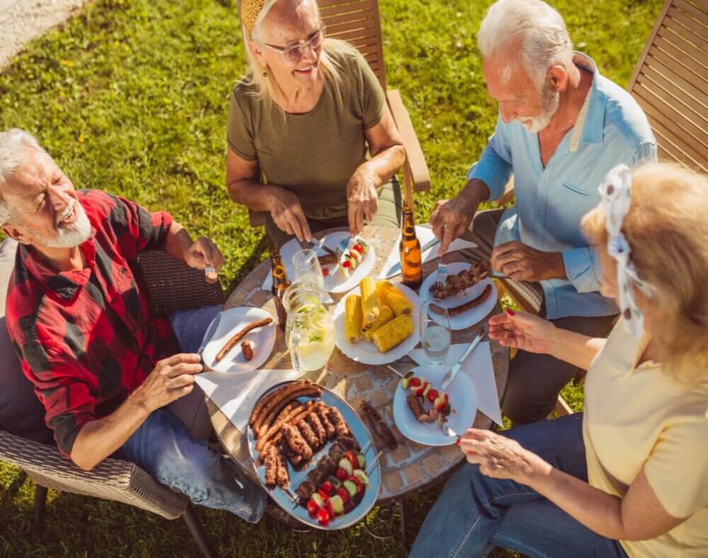 Four elderly people enjoying a meal together at an outdoor table, with various dishes and drinks in a sunny setting. - Home Instead Poole
