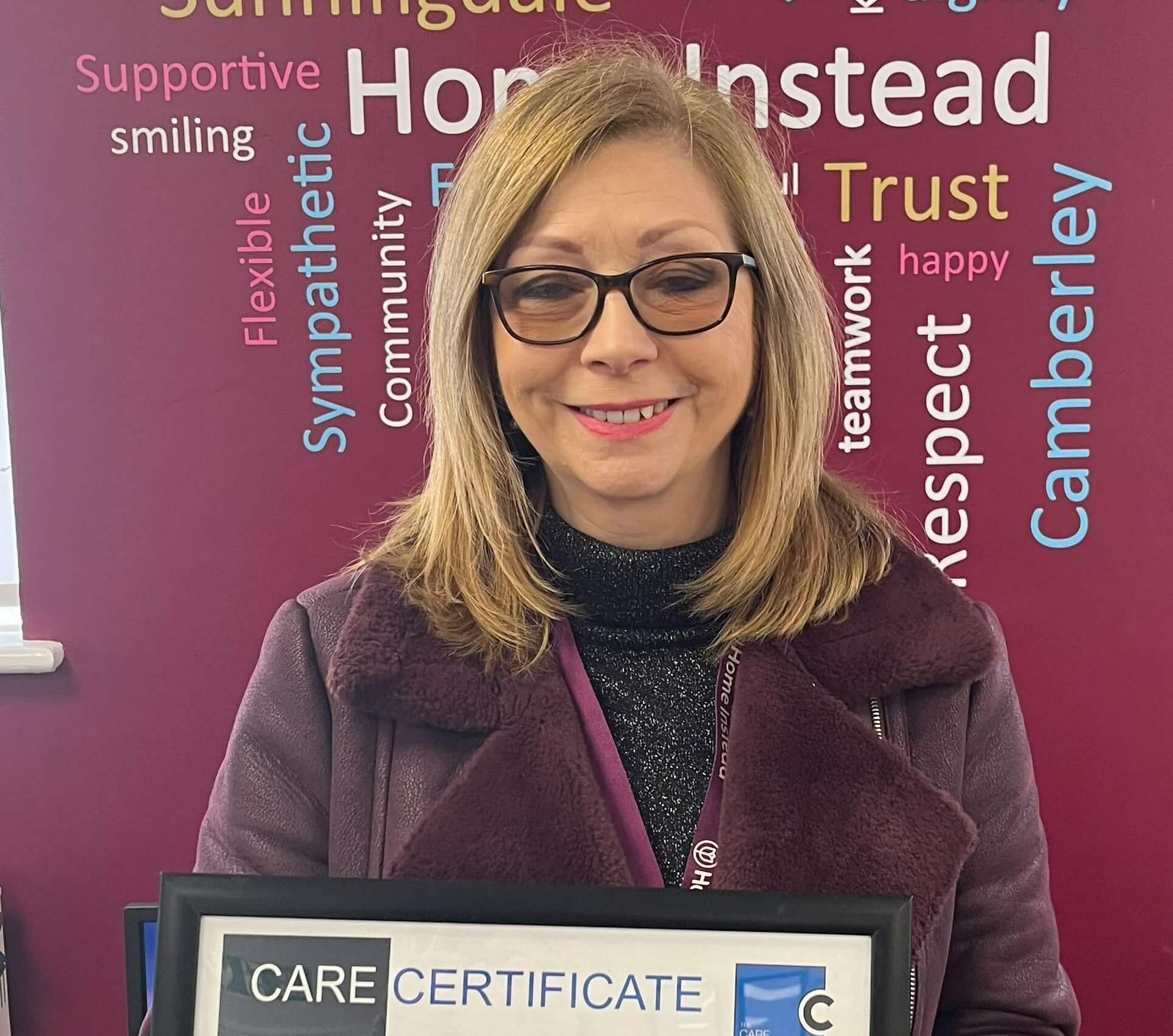 A woman smiling and holding a framed Care Certificate, standing against a colorful word cloud background. - Home Instead