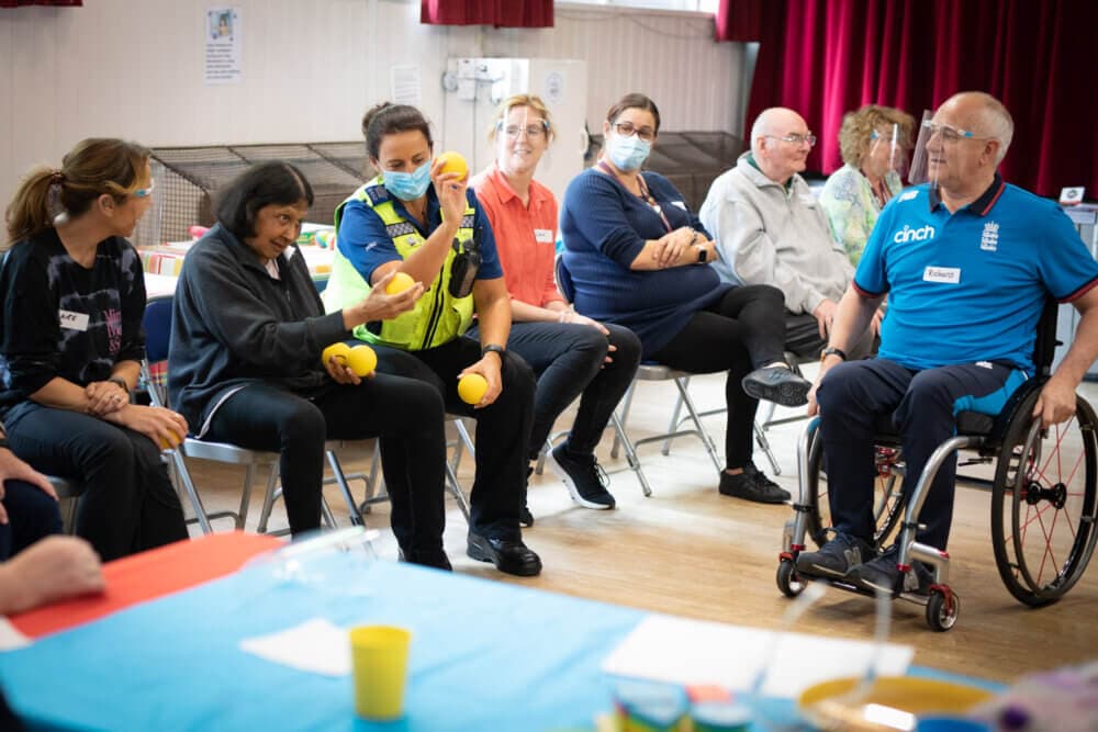 A group of people, some wearing masks, sit in a circle while a woman in a safety vest shows items to the group. - Home Instead