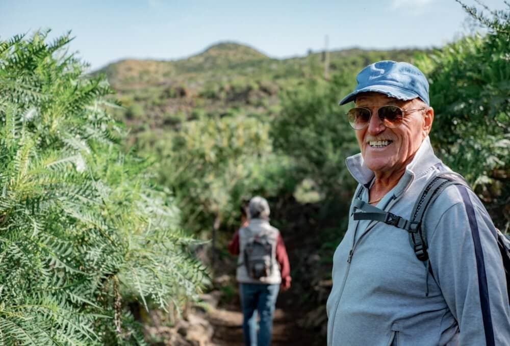 An older man wearing a blue cap and sunglasses smiles while hiking on a trail, with another hiker ahead in the background. - Home Instead