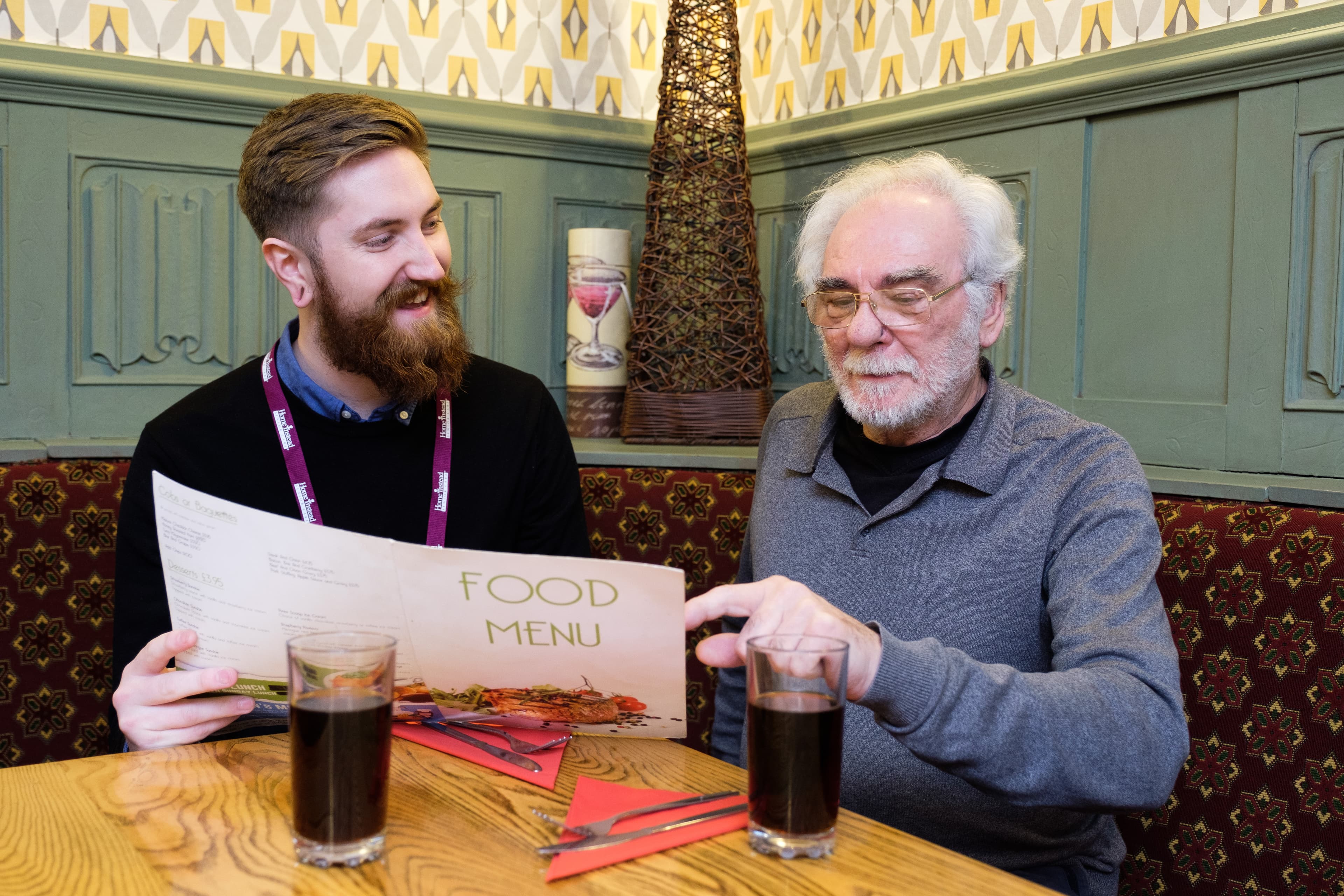 Two men sitting at a restaurant table, looking at a menu together, with two glasses of soda in front of them. - Home Instead