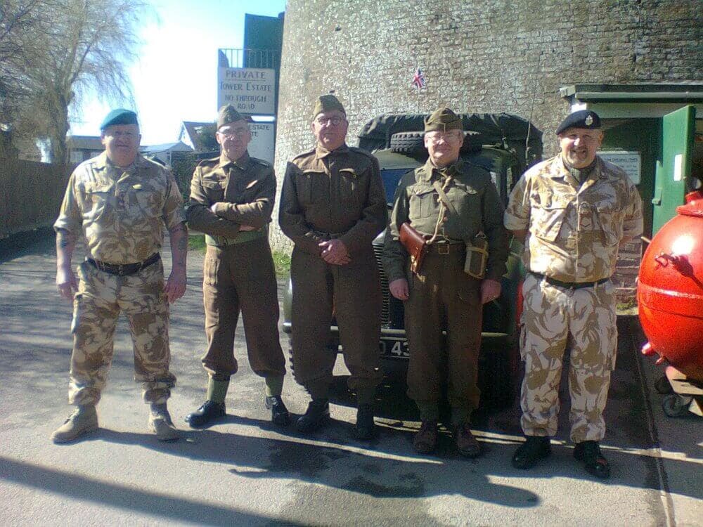 Five men dressed in military uniforms stand in front of a vintage military vehicle outside of a brick building. - Home Instead