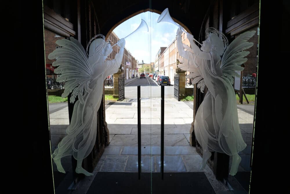 Frosted glass doors with etched angels blowing trumpets, opening to a street with cars and buildings. - Home Instead
