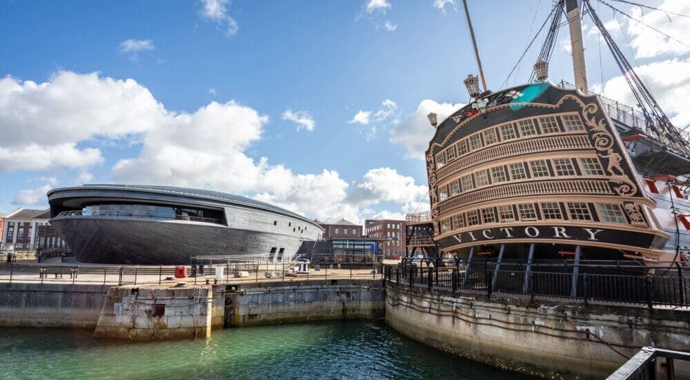 Modern museum building and the HMS Victory, an 18th-century ship, docked at Portsmouth Historic Dockyard under a blue sky. - Home Instead