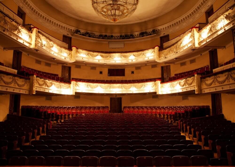An empty, ornate theatre auditorium with red and gold decor, chandelier, and balcony seating, illuminated warmly. - Home Instead Southampton