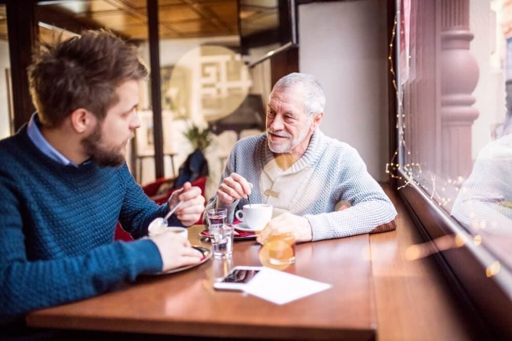 Two men, one younger and one older, sitting at a café table, having a conversation over drinks and desserts. - Home Instead Southampton