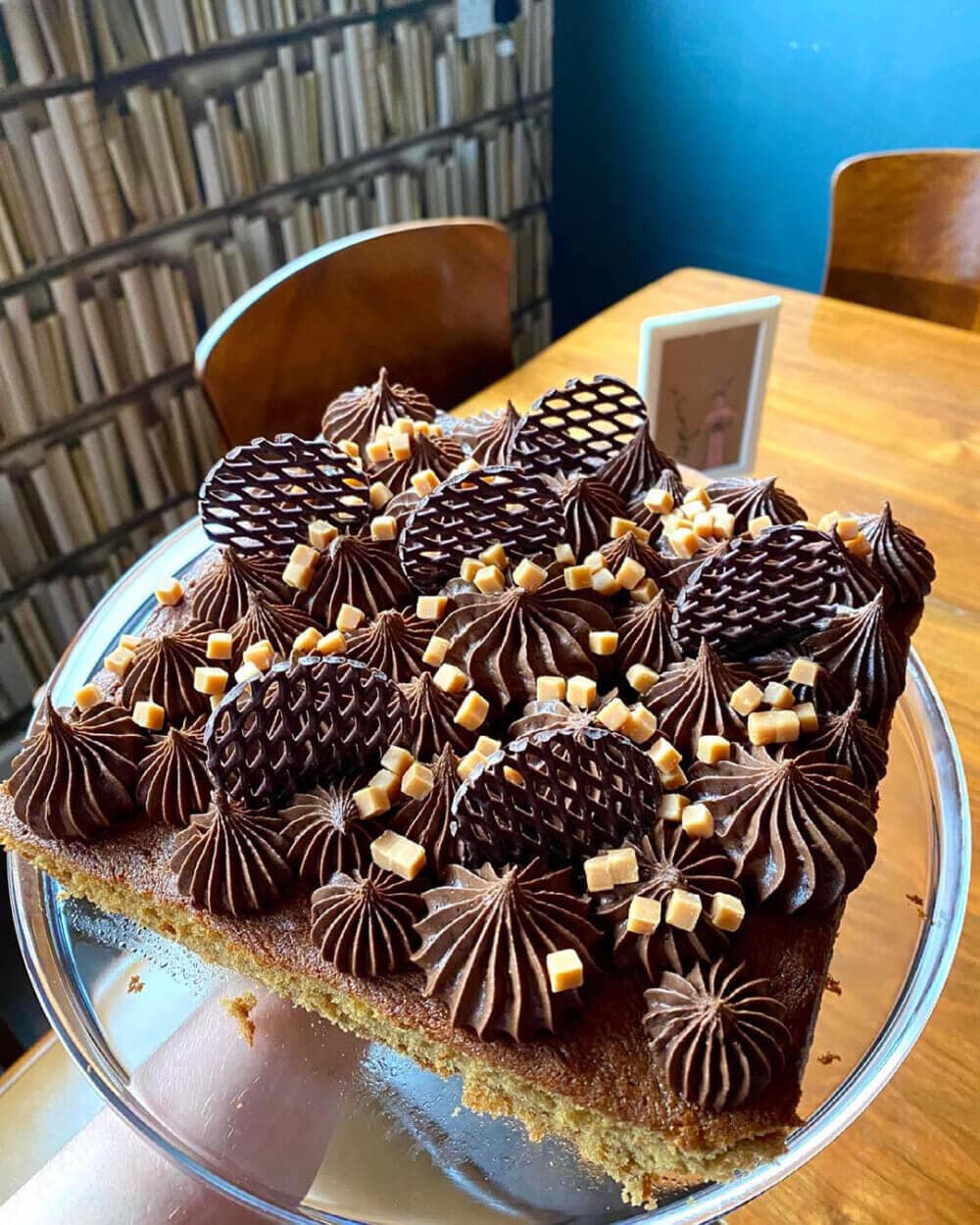 A square chocolate cake topped with chocolate frosting swirls, chocolate pieces, and small caramel cubes on a glass plate. - Home Instead