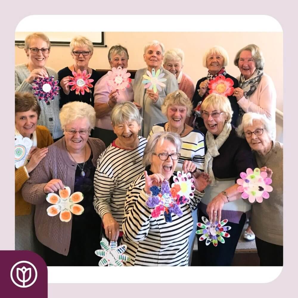 A group of elderly people holding colorful paper flowers and smiling together, standing in a room. - Home Instead