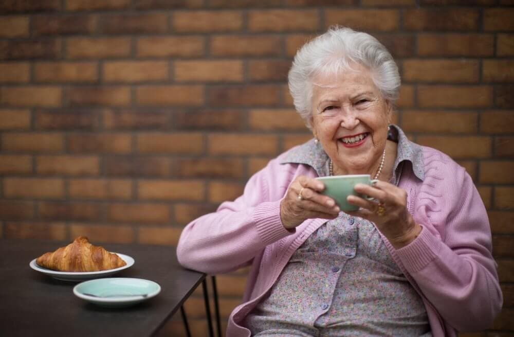 Elderly woman with white hair smiling, holding a cup, sitting at a table with a croissant in front of a brick wall. - Home Instead