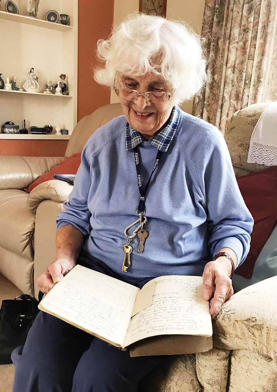 Elderly woman with white hair and glasses reads from an open notebook while sitting on a beige couch at home. - Home Instead
