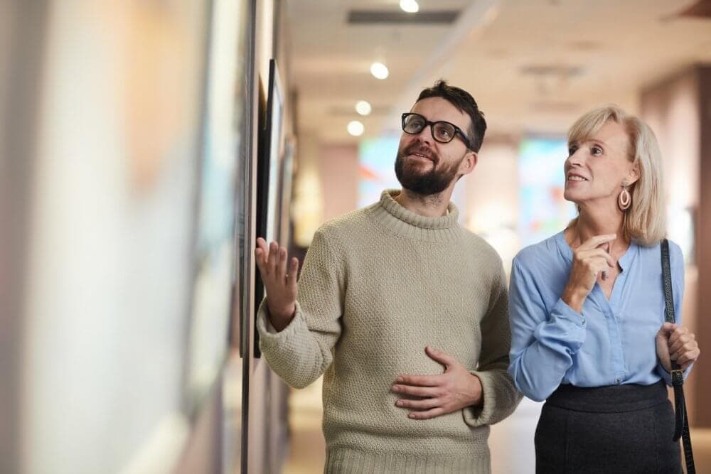 Two people enjoying an art exhibit; a man on the left points at a painting while a woman on the right looks on attentively. - Home Instead
