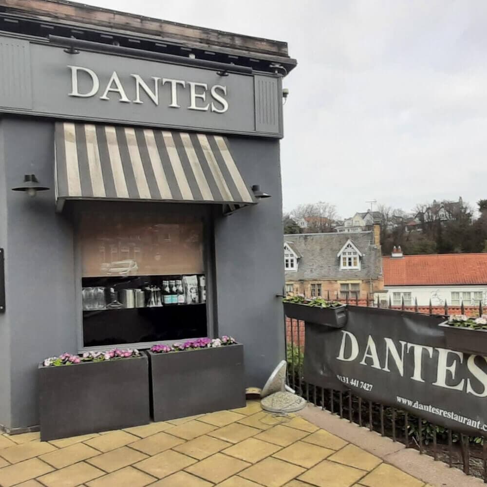 Small gray building with a striped awning displaying the name "Dantes." A railing with a "DANTES" banner is in front. - Home Instead
