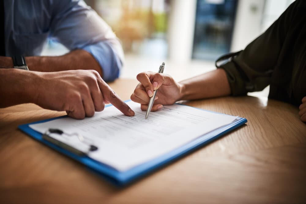 Two people review and sign a document on a clipboard at a wooden table, focusing on the pen and paper details. - Home Instead