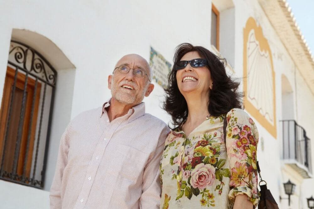 A smiling man and woman wearing sunglasses stand outside a white building with wooden doors. - Home Instead