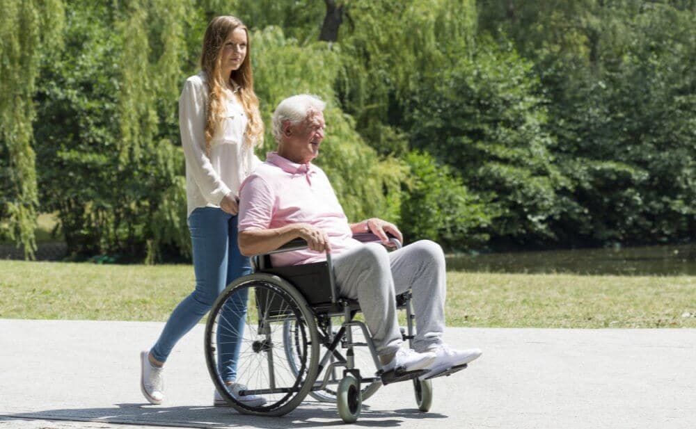 A young woman pushes an older man in a wheelchair along a park path on a sunny day. - Home Instead