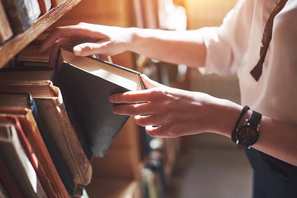 Person in a white shirt pulling a book from a library shelf, lit by warm natural light. - Home Instead