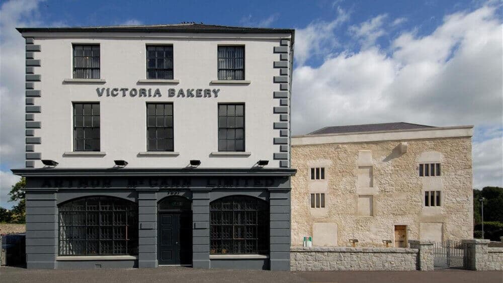 A historic building labeled "Victoria Bakery" stands beside a stone-walled structure under a partly cloudy sky. - Home Instead