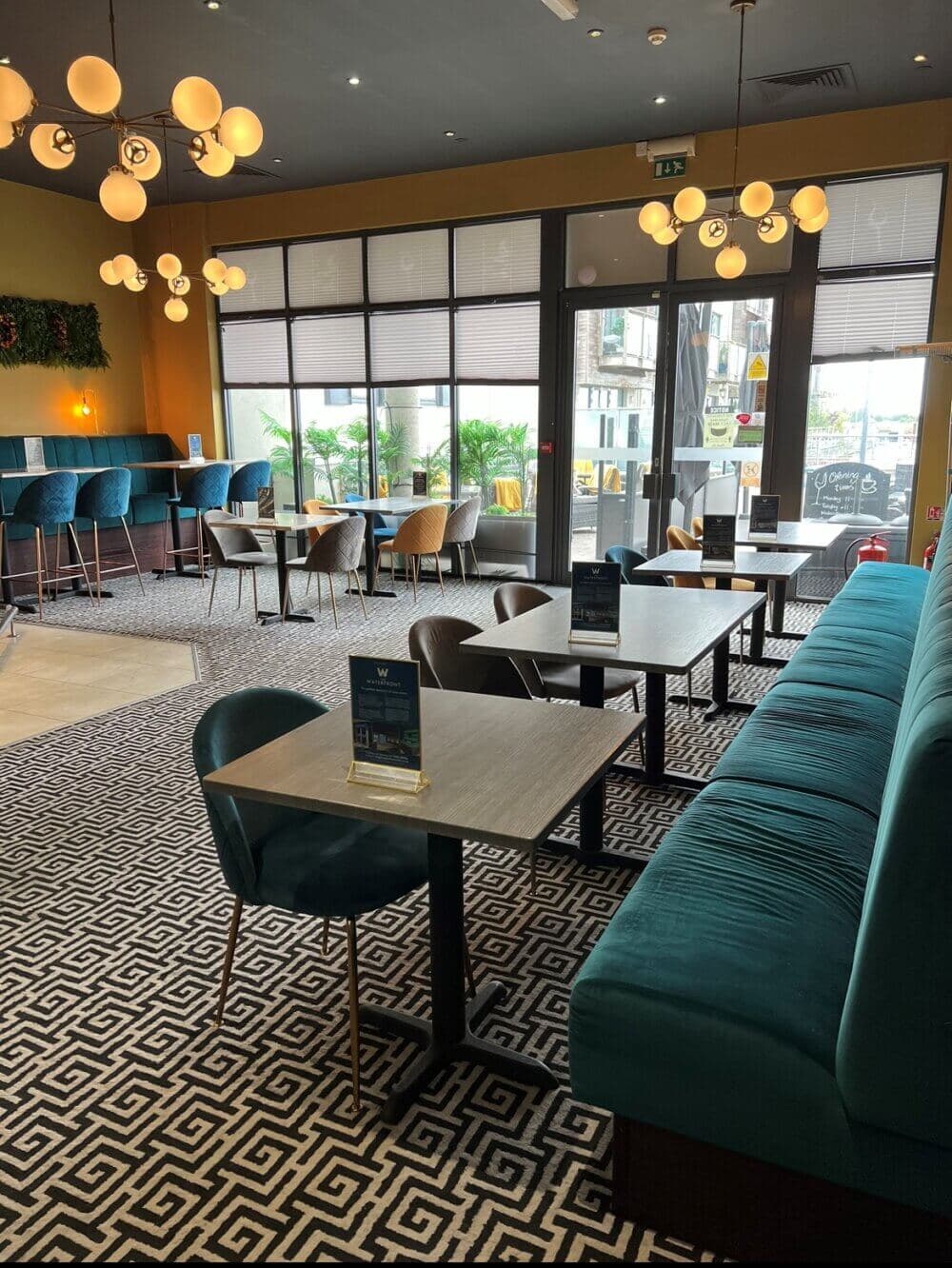 A modern café with teal velvet chairs, patterned carpet, and round pendant lights illuminating the chic, empty space. - Home Instead