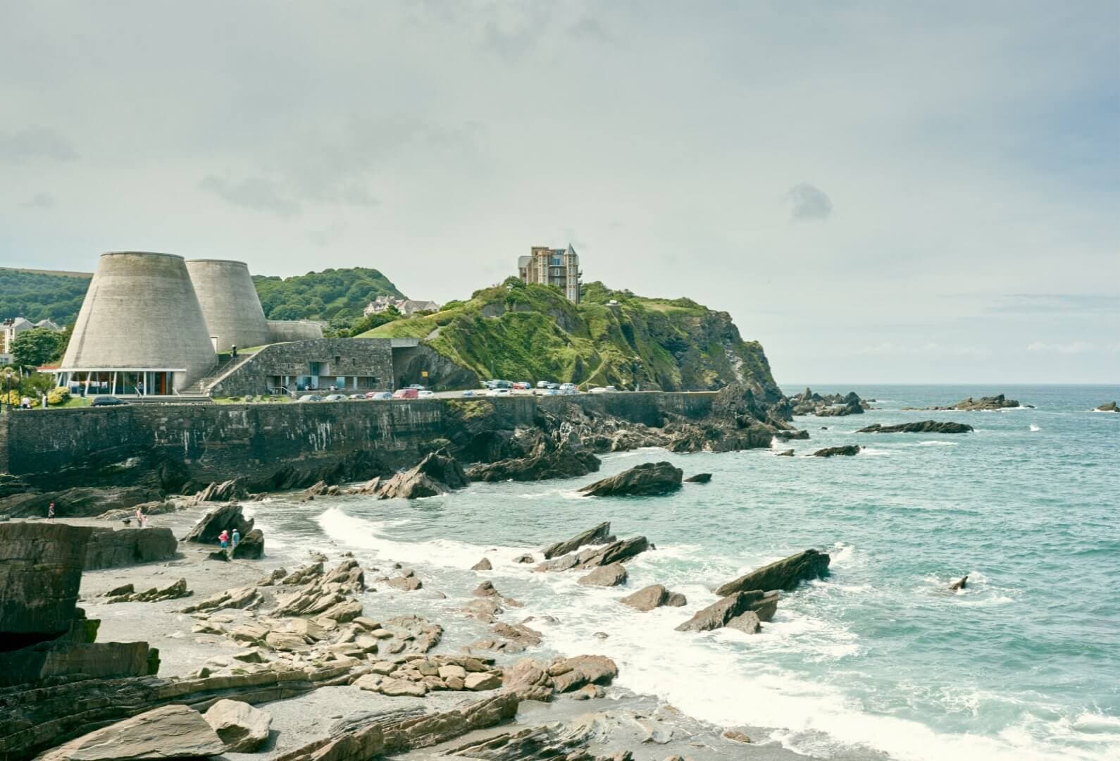 Coastal view with modern buildings on the left, a rocky shoreline, and a distant building on a green hill. - Home Instead