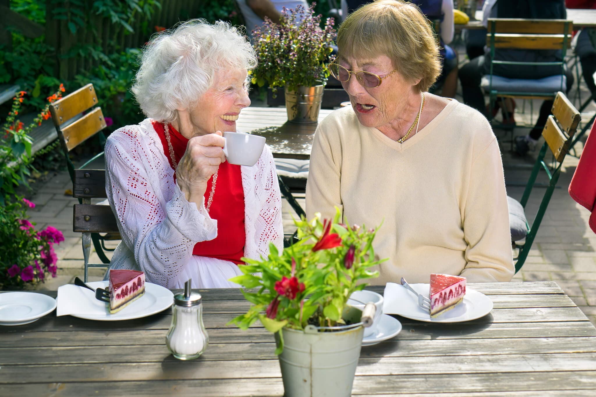 Two elderly women smiling, sitting at an outdoor café table with tea and cake, surrounded by plants and flowers. - Home Instead