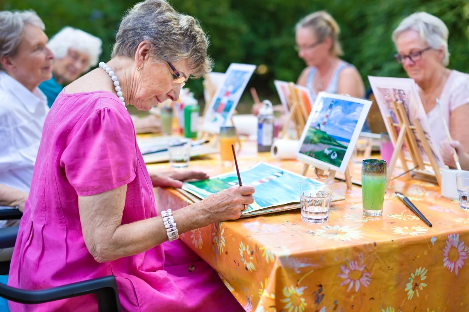 Elderly women sit at a table outdoors, engaging in a painting activity, each with their own canvas and materials. - Home Instead