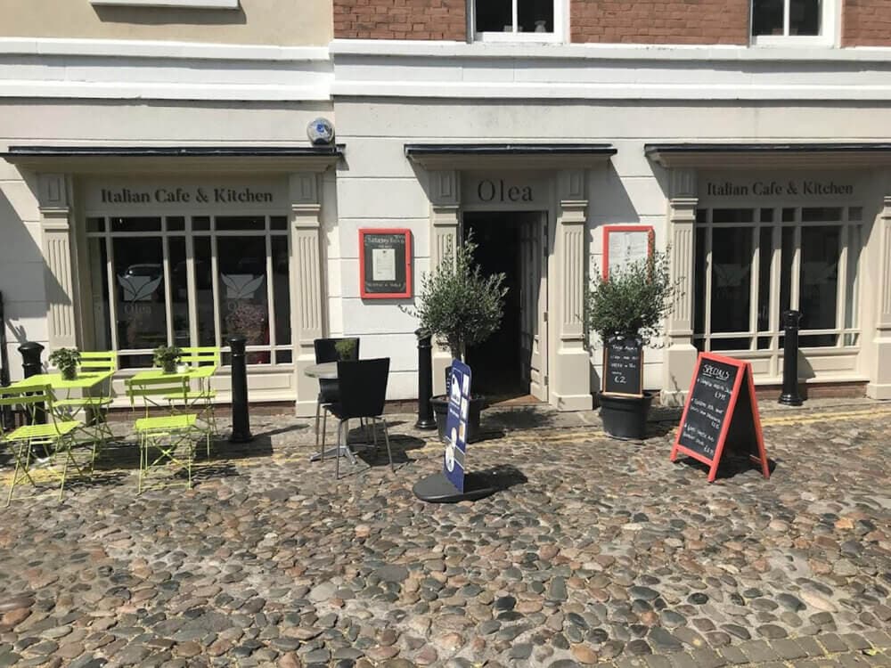Front of the "Olea" Italian Café & Kitchen with outdoor seating and menu boards on a cobblestone street. - Home Instead