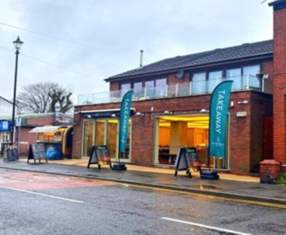 Street view of takeaway restaurant with green signs, glass windows, and a lit interior on a rainy day. - Home Instead