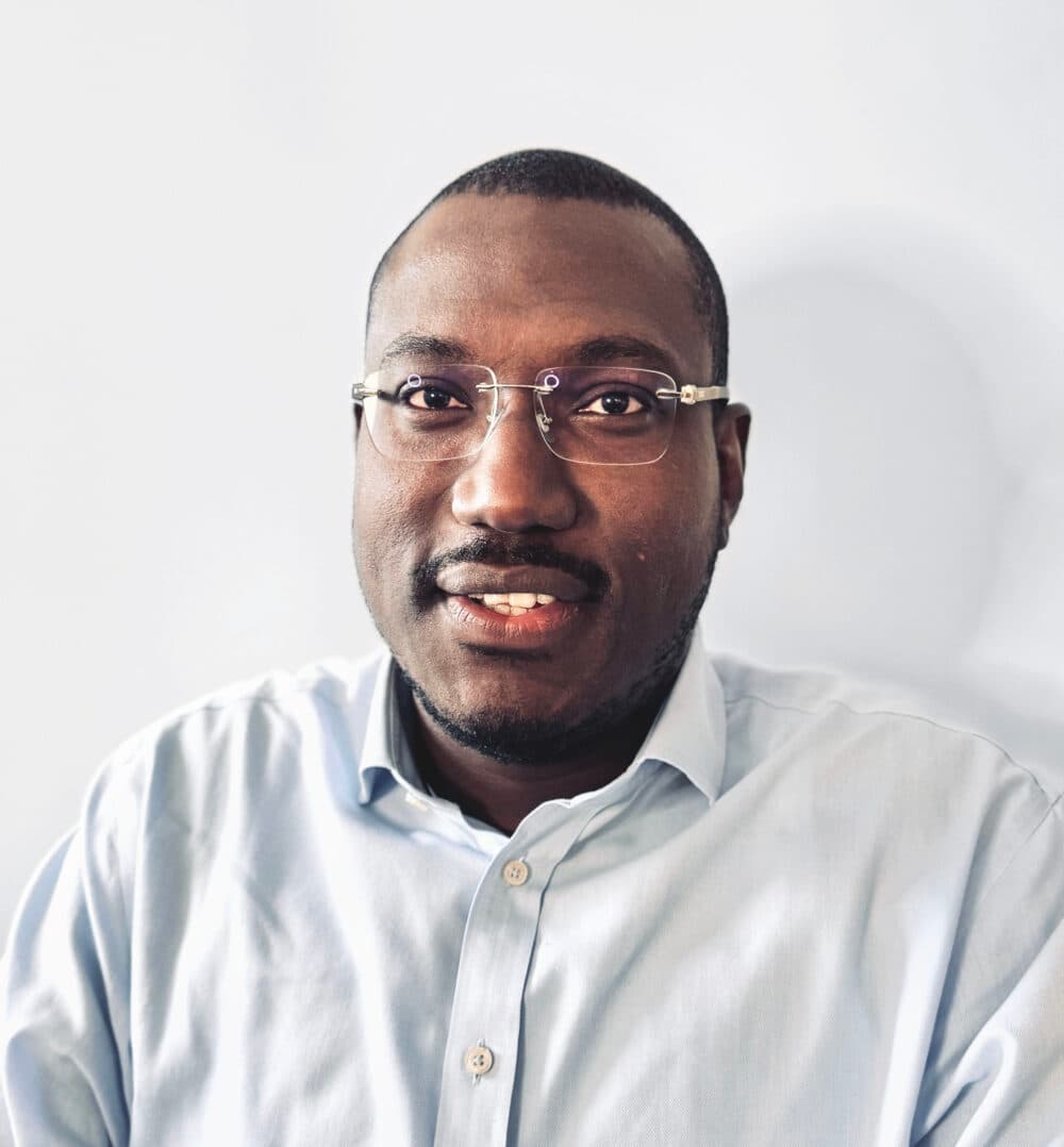 A man wearing glasses and a light blue collared shirt smiles at the camera against a plain background. - Home Instead